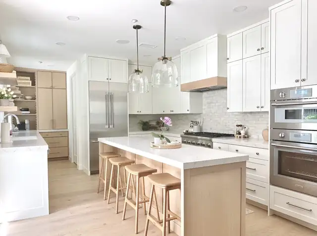 White shaker kitchen with a long quartz island, light oak counter stools, glass pendants and double wall ovens