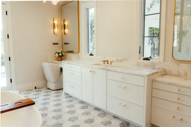 Bathroom remodel with a long white vanity, brass sconces, a gold-framed mirror and a patterned tile floor
